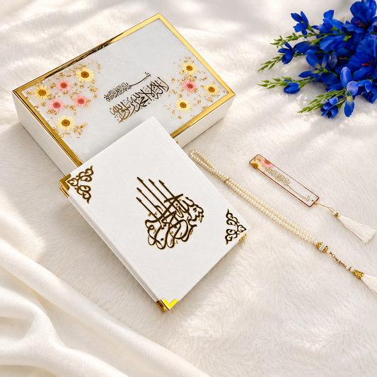 White Islamic prayer book set with gold foil calligraphy, a matching decorative gift box with floral print, a pearl tassel bookmark, and blue flowers on a soft white fabric background.