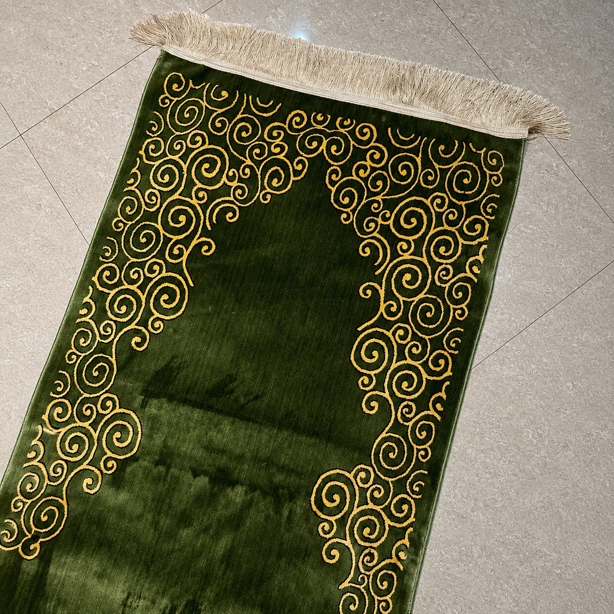 A green velvet prayer rug with gold embroidered swirl patterns and a beige fringed edge, laid diagonally on a light gray tiled floor.