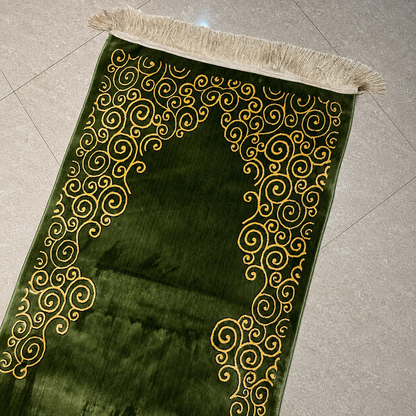A green velvet prayer rug with gold embroidered swirl patterns and a beige fringed edge, laid diagonally on a light gray tiled floor.