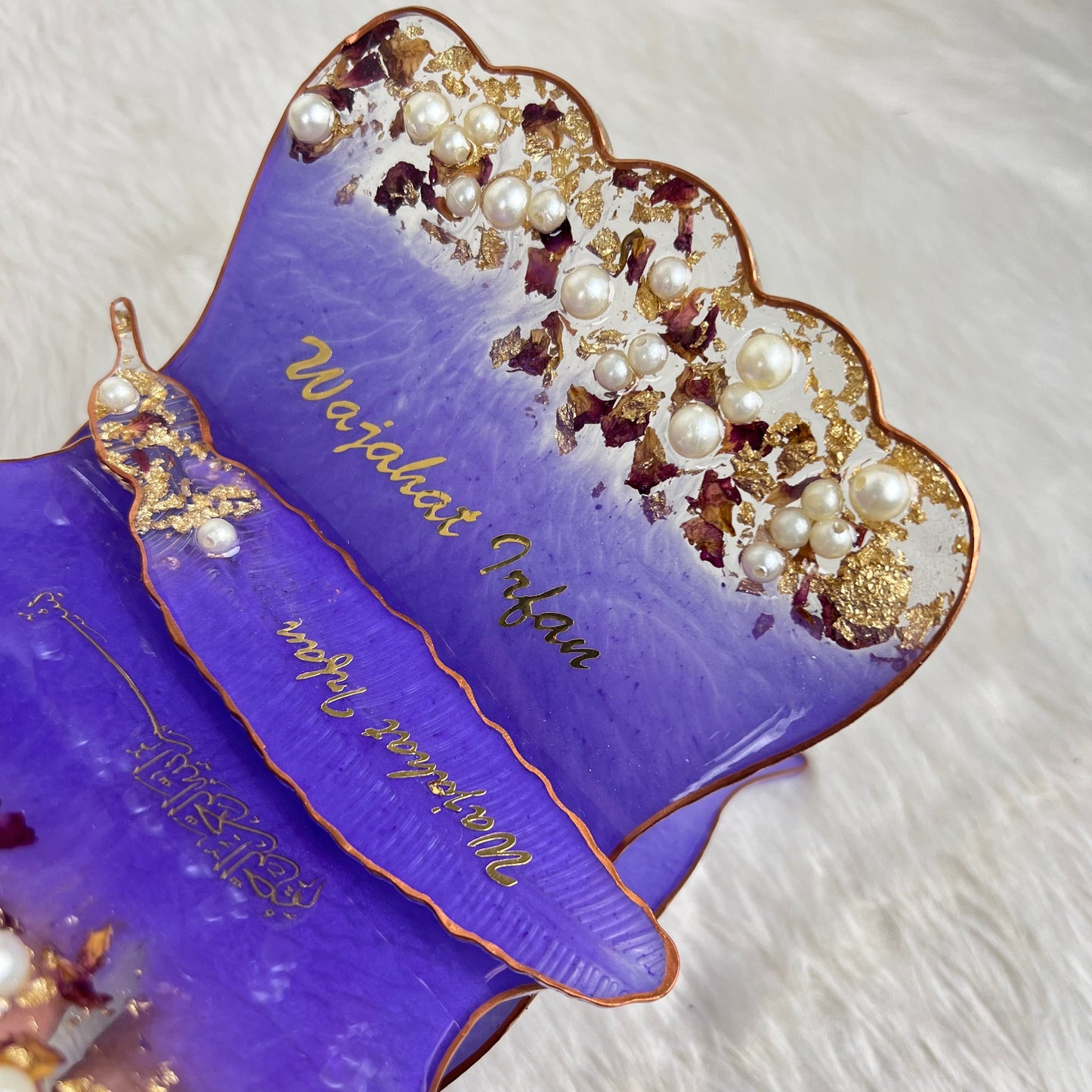 A close-up of a purple decorative tray with scalloped edges, gold script writing, gold trim, and embellishments of faux pearls and gold leaf, resting on a white faux fur surface.