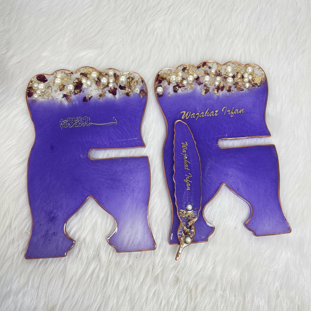 Two purple enamel Islamic wall art plaques shaped like the letter 'R' with gold Arabic calligraphy, pearl embellishments, and a miniature feather pen, displayed on a white faux fur background.