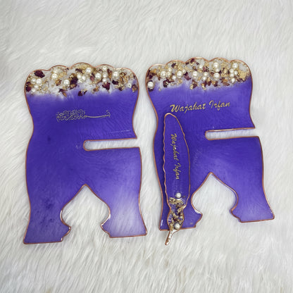 Two purple enamel Islamic wall art plaques shaped like the letter 'R' with gold Arabic calligraphy, pearl embellishments, and a miniature feather pen, displayed on a white faux fur background.