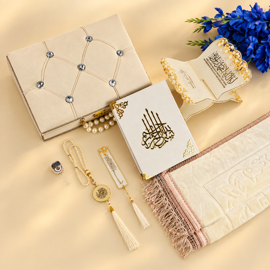 A styled flat-lay of Islamic prayer accessories and a matching gift set on a warm beige background. The set includes a cream-colored prayer rug with pink and gold stripes, a white Quran with gold Arabic calligraphy, a cream prayer mat, a cream prayer beads necklace with gold and pearl details, a cream prayer mat with a tassel, a cream decorative box with silver rhinestones, and a small cream prayer card holder, all arranged with blue flowers.