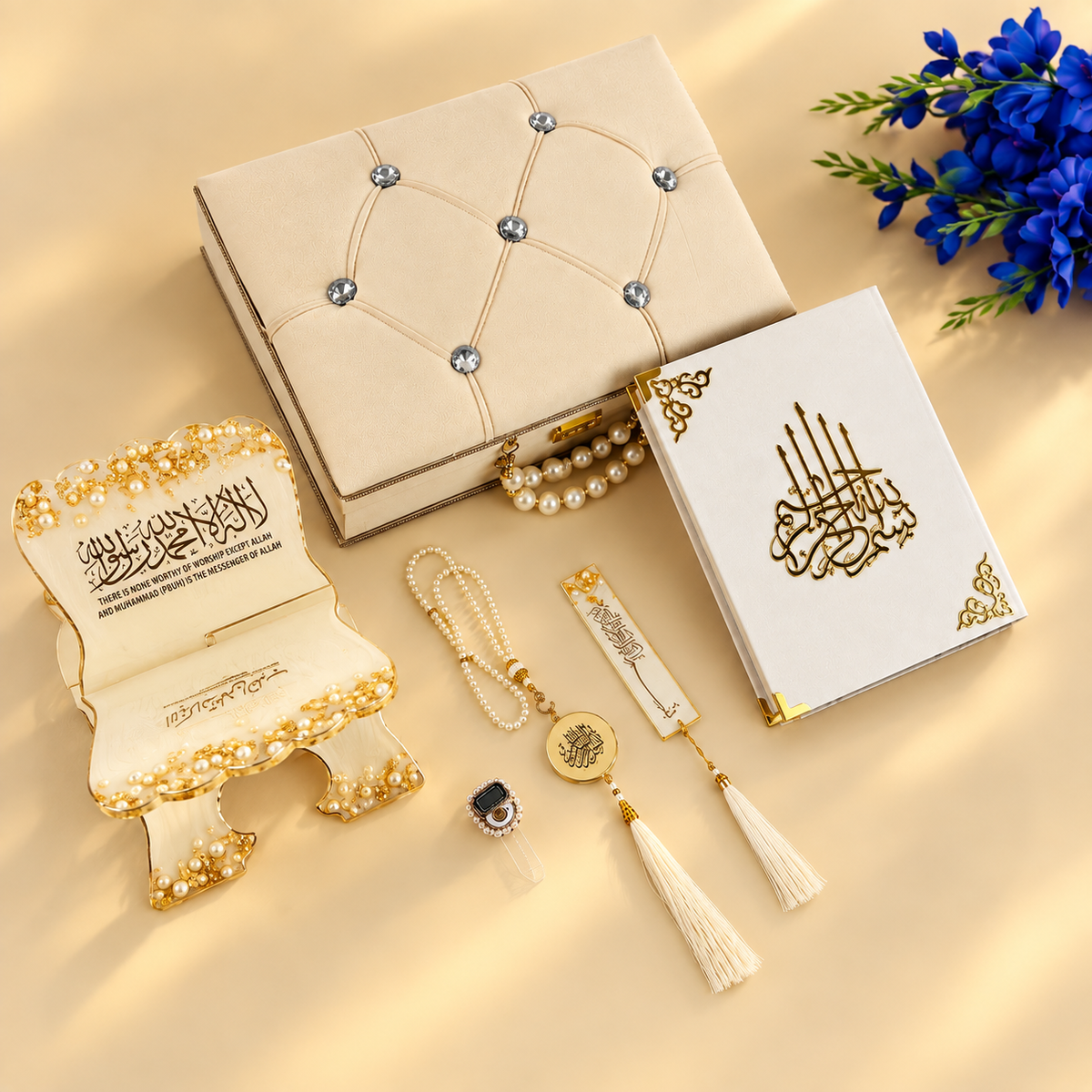 A styled flat-lay of Islamic prayer accessories on a warm beige background, including a cream and gold Quran with Arabic calligraphy, a matching small stand, pearl bead necklaces, a tassel with a circular medallion, a small rectangular tag, and a decorative box with rhinestone tufting. Blue flowers appear in the top right corner.