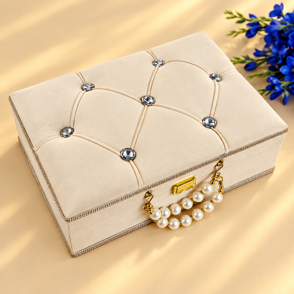 A cream-colored velvet jewelry box with tufted diamond stitching, crystal-like rhinestone accents, a gold-tone clasp, and a pearl-trimmed handle, shown on a warm beige background with blue flowers in the corner.