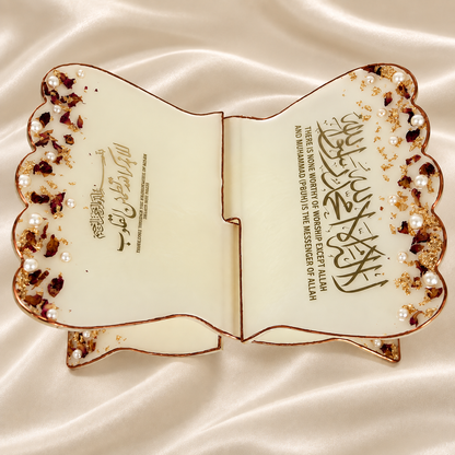 A premium resin rehal displayed open on a cream satin background, featuring elegant gold Arabic calligraphy, delicate rose petals, pearl embellishments, gold flakes, and a metallic border. The image highlights its elegant design, beautiful calligraphy, sturdy construction, and premium resin finish.