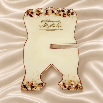 A premium resin rehal displayed open on a cream satin background, featuring elegant gold Arabic calligraphy, delicate rose petals, pearl embellishments, gold flakes, and a metallic border. The image highlights its elegant design, beautiful calligraphy, sturdy construction, and premium resin finish.