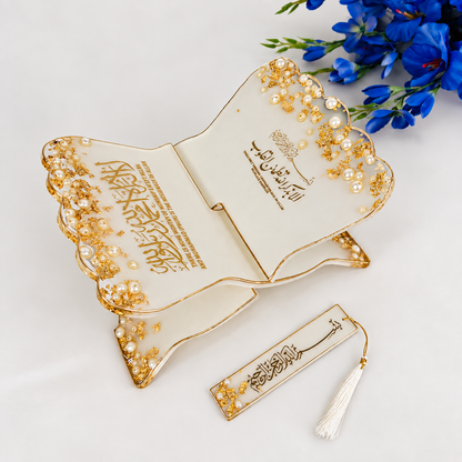 An ornate open book-shaped decorative plaque or tray in warm ivory with gold trim, pearl-like embellishments, and Arabic calligraphy, shown on a white surface with blue flowers in the corner and a matching bookmark-like tag with a tassel below.