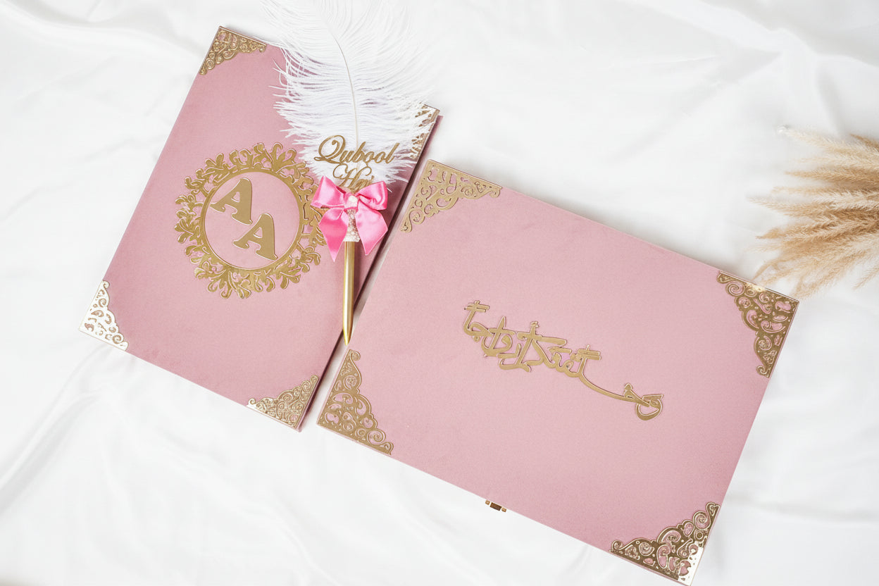 Pink invitation cards with gold text and a feather pen on a white surface