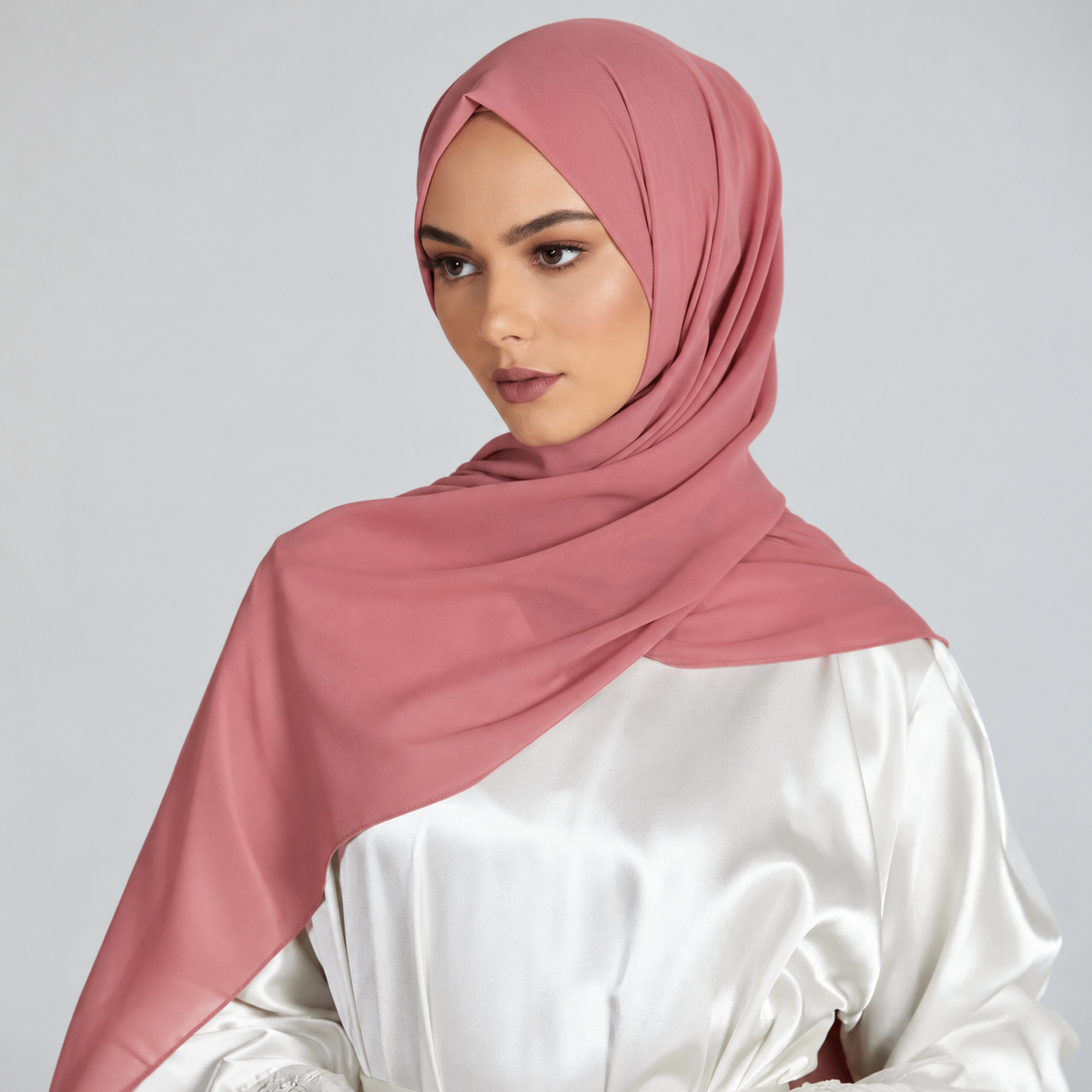 A woman wearing a dusty rose pink hijab and a white satin top, photographed against a neutral light gray studio background.