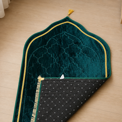 A dark teal velvet prayer rug with gold trim and a black patterned underside, laid out on a light wood floor.