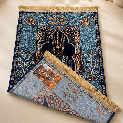 A rectangular patterned prayer rug laid on a light beige floor, with one corner folded back to show the reverse side. The rug features an ornate blue and navy central motif with floral borders and gold fringe.

Designed for those who appreciate premium Islamic aesthetics and traditional craftsmanship, this prayer rug is perfect for everyday worship as well as meaningful gifting.