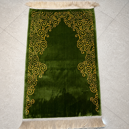 A green velvet prayer rug with gold embroidered scrollwork and cream fringe, laid flat on a light gray tiled floor.