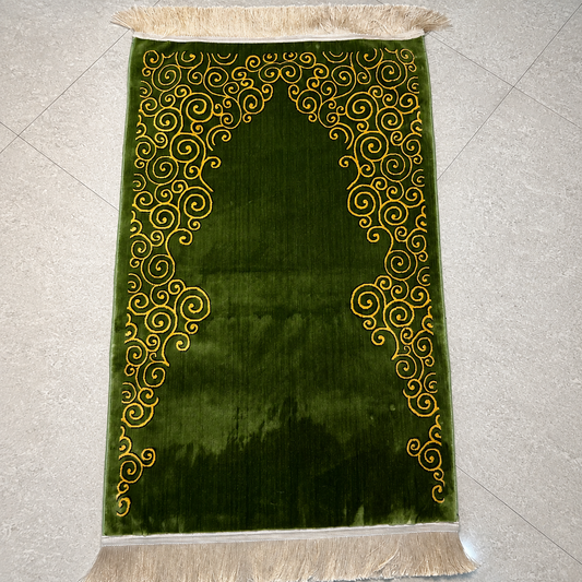 A green velvet prayer rug with gold embroidered scrollwork and cream fringe, laid flat on a light gray tiled floor.