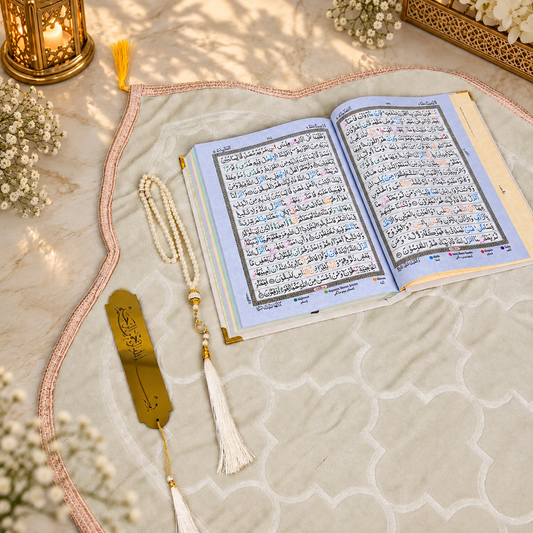An open Quran with Arabic script on light blue pages, laid on a beige patterned prayer mat with a pink border. A white beaded tassel necklace, a gold bookmark, and small white flowers are arranged around it, with warm sunlight and shadows across the scene.