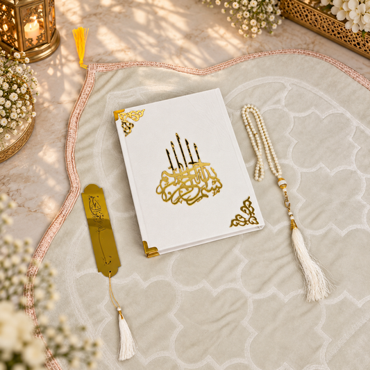 A white Quran with ornate gold Arabic calligraphy on the cover, styled on a beige patterned prayer mat with a gold tassel bookmark, a white pearl rosary, and decorative floral accents.