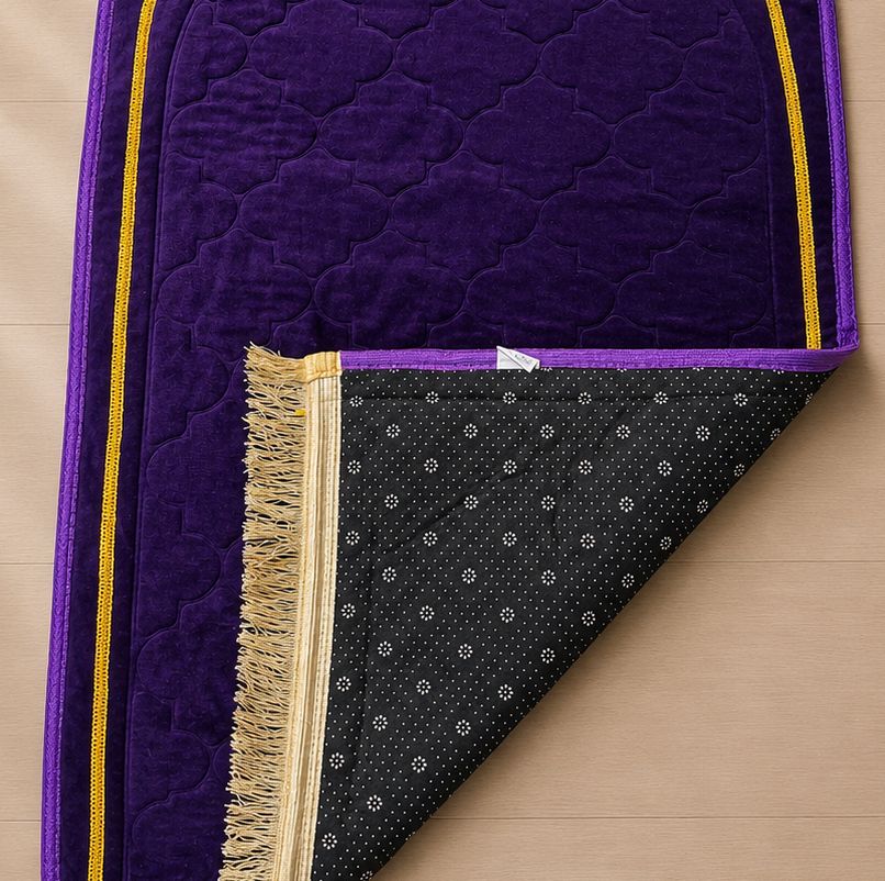 A purple prayer rug with gold trim and a black patterned underside with fringe, laid on a light wood floor next to a potted plant.