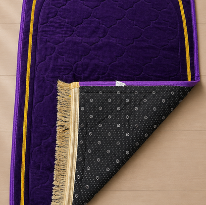 A purple prayer rug with gold trim and a black patterned underside with fringe, laid on a light wood floor next to a potted plant.