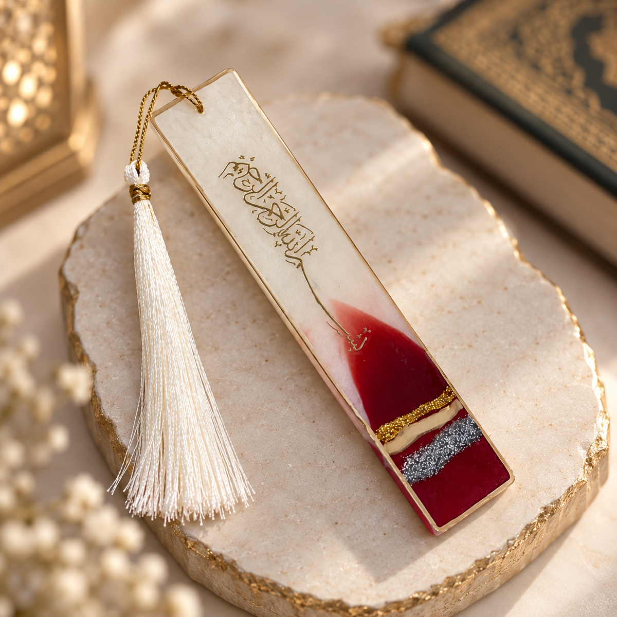 A rectangular resin bookmark with Arabic calligraphy, featuring a red and silver geometric design at the bottom, a white tassel, and a gold loop, resting on a decorative stone slab.