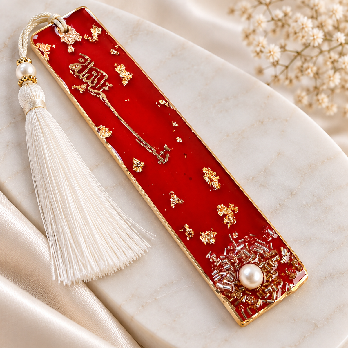 A rectangular red and gold bookmark with Arabic calligraphy, a white tassel, and a pearl accent, displayed on a cream marble surface with dried flowers.