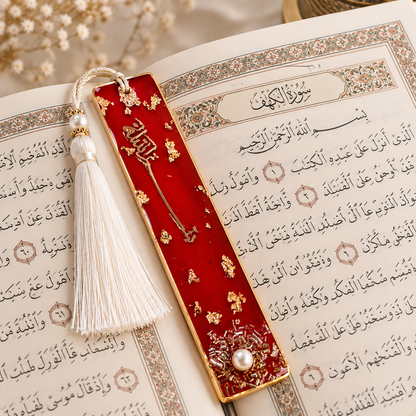 A red velvet Quran bookmark with gold foil details, a white tassel, and a pearl accent, resting on an open book with Arabic script.