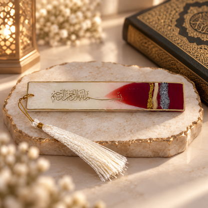 A rectangular Islamic bookmark with Arabic calligraphy, featuring a cream background, a deep red gradient section, gold and silver stripes, and a white tassel, displayed on a stone slab with a book and lantern in the background.
