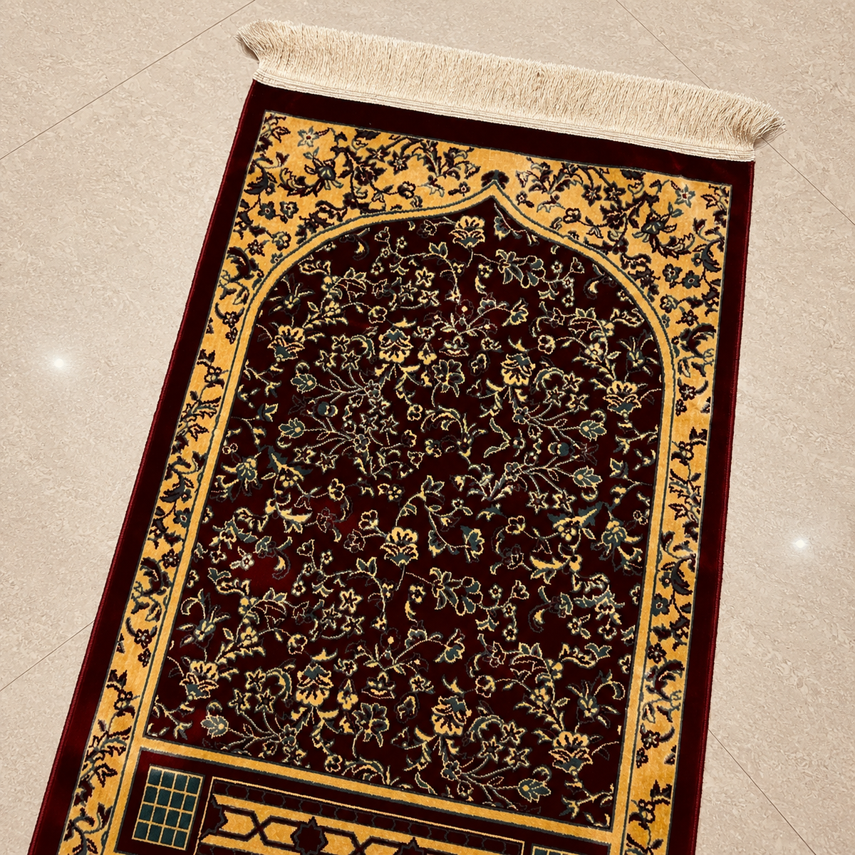 A rectangular prayer rug with an ornate floral pattern, featuring a deep burgundy center, a golden-yellow border, and cream fringe at the top, laid out on a light beige tiled floor.