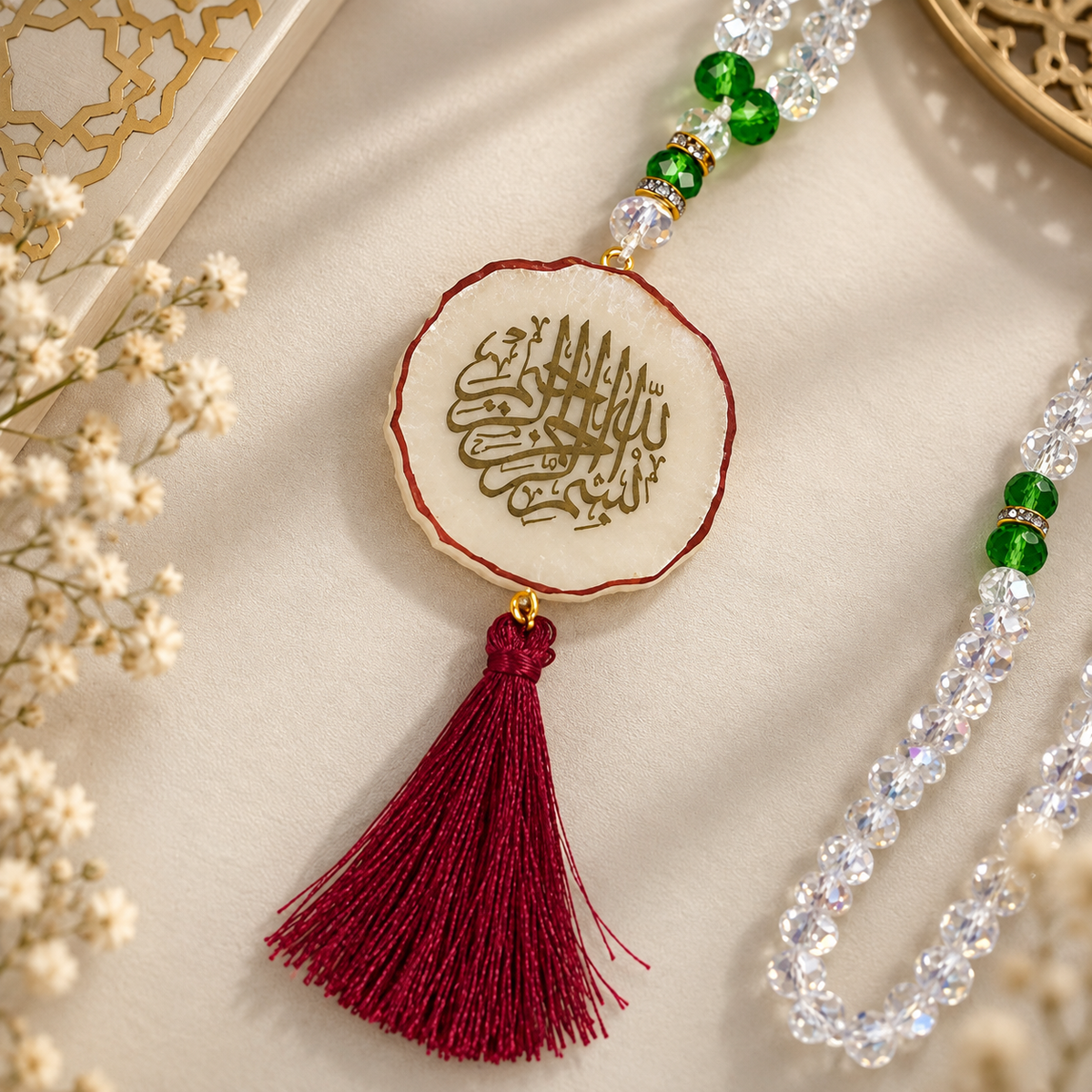 A decorative Islamic prayer bead necklace (Tasbih) featuring a round beige wooden medallion with gold Arabic calligraphy, a deep red tassel, and green and clear faceted beads, styled on a beige fabric background with dried flowers.