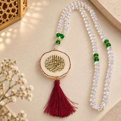 A beaded necklace with clear faceted beads, green accent beads, and a central round white pendant featuring gold Arabic calligraphy, finished with a deep red tassel. The necklace is styled on a beige surface with soft shadows and small dried flowers.