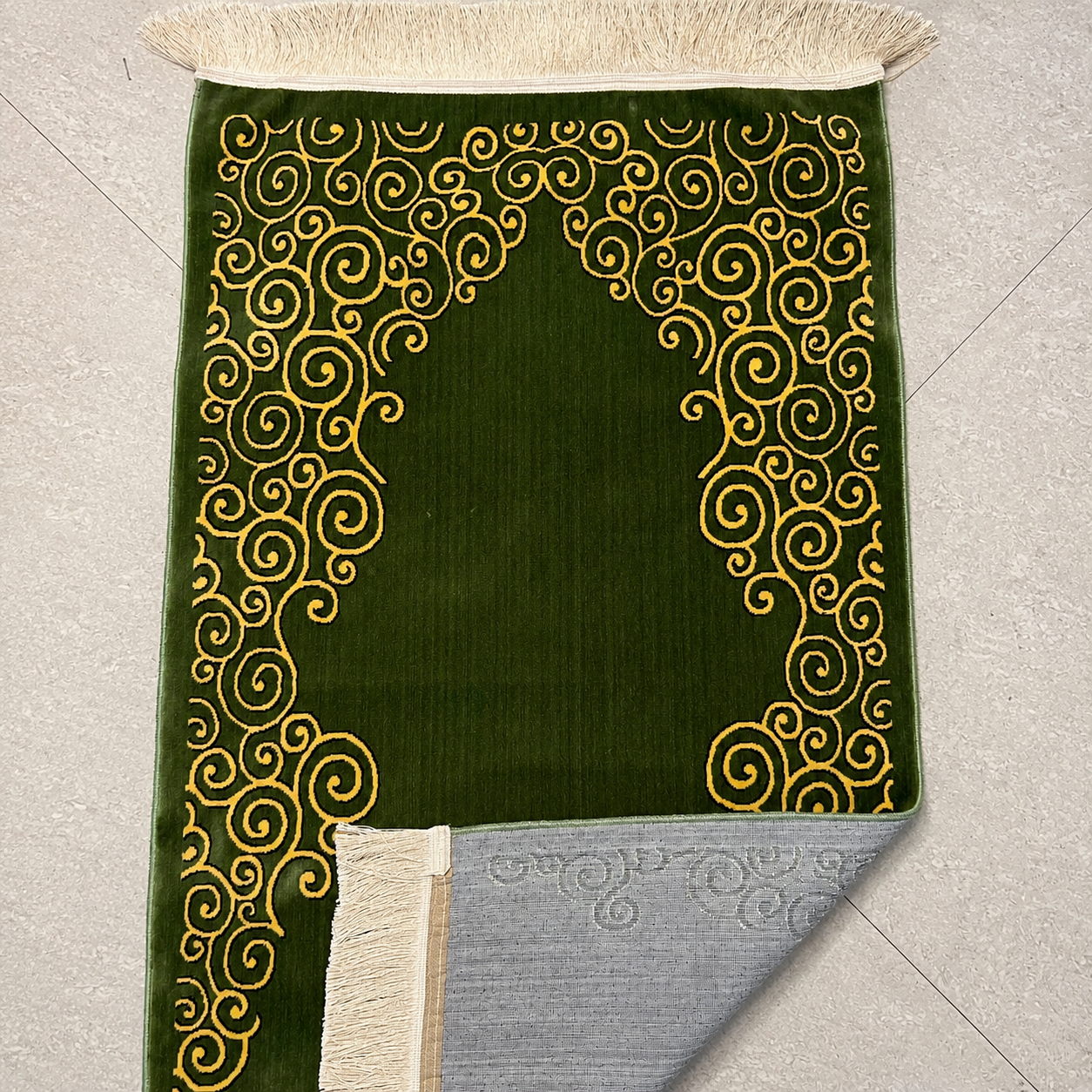 A green prayer rug with gold scrollwork patterns and cream fringe, shown laid flat on a light gray tiled floor with one corner folded over to reveal the backing.