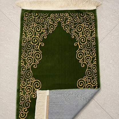 A green prayer rug with gold scrollwork patterns and cream fringe, shown laid flat on a light gray tiled floor with one corner folded over to reveal the backing.