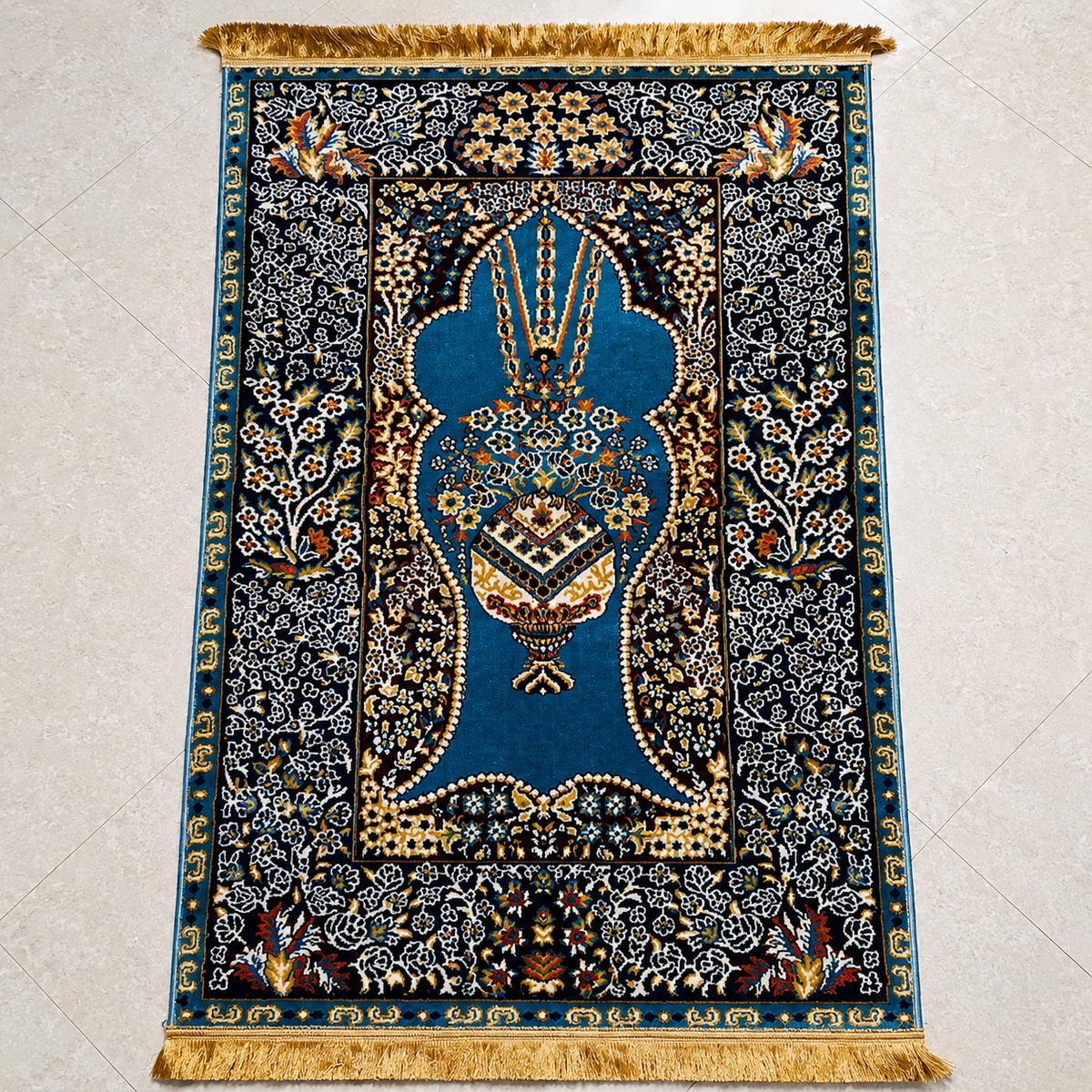 An ornate rectangular rug with a central blue medallion and intricate floral motifs in gold, beige, and black, finished with a gold fringe at the top and bottom, photographed flat on a light beige floor.