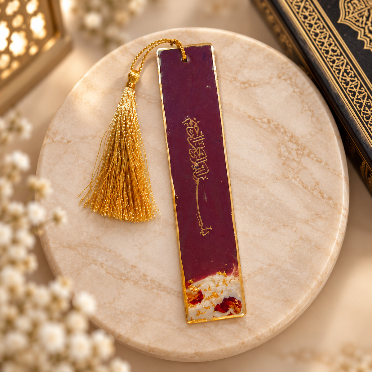A rectangular maroon bookmark with gold trim and a gold tassel, featuring gold Arabic calligraphy and a small floral design near the bottom, displayed on a round beige marble coaster.