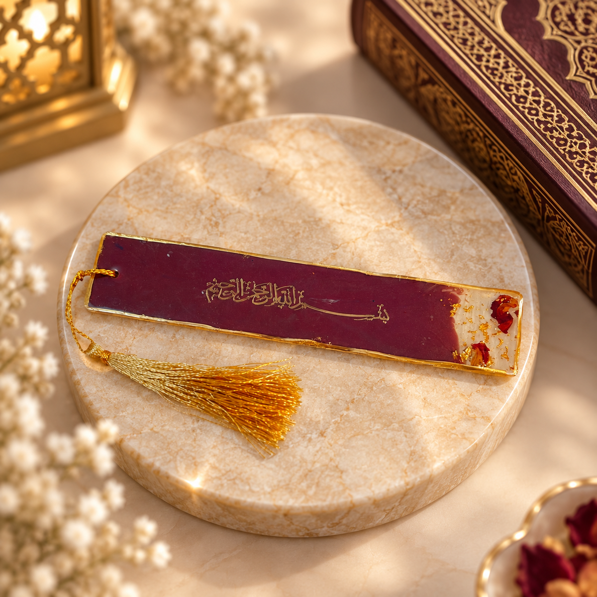 A decorative book bookmark with a gold tassel, featuring a deep burgundy-red center with gold Arabic calligraphy and dried red flower accents, displayed on a round beige marble coaster.
