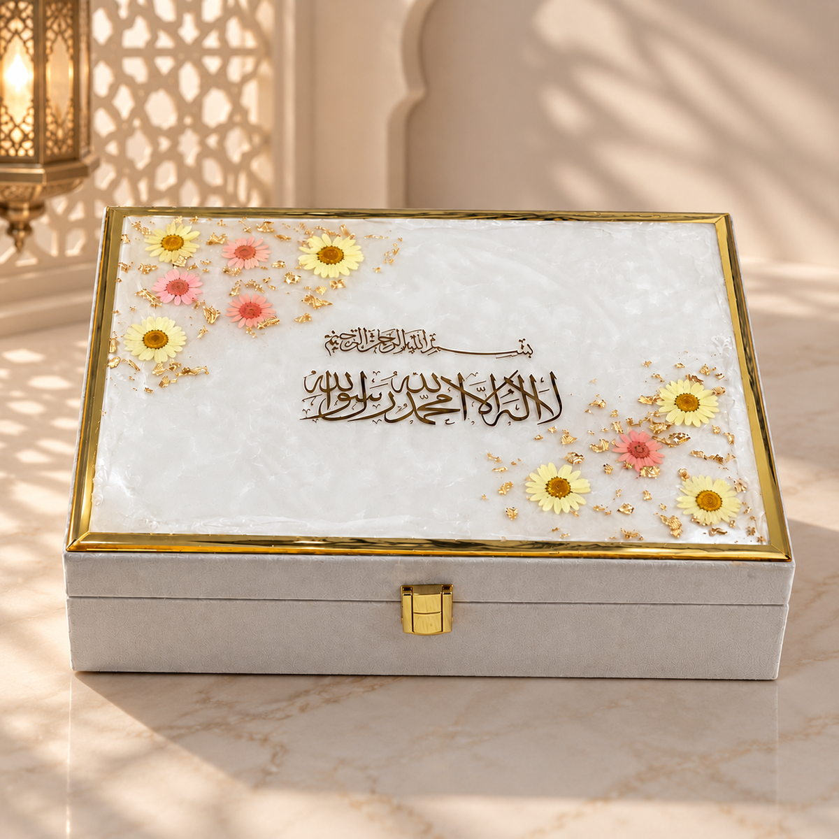 A rectangular decorative jewelry box with a white marbled lid featuring gold Arabic calligraphy, small yellow and pink flowers, and a gold border, set on a marble surface with warm ambient light.