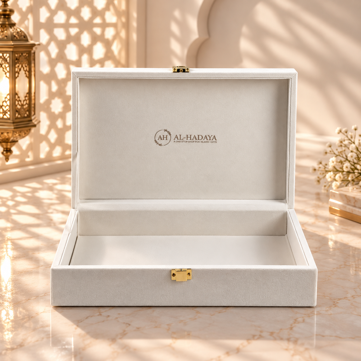 An open white luxury presentation box with a gold clasp and the brand name 'AL-HADAYA' printed inside the lid, set on a marble surface with warm sunlight and shadow patterns.