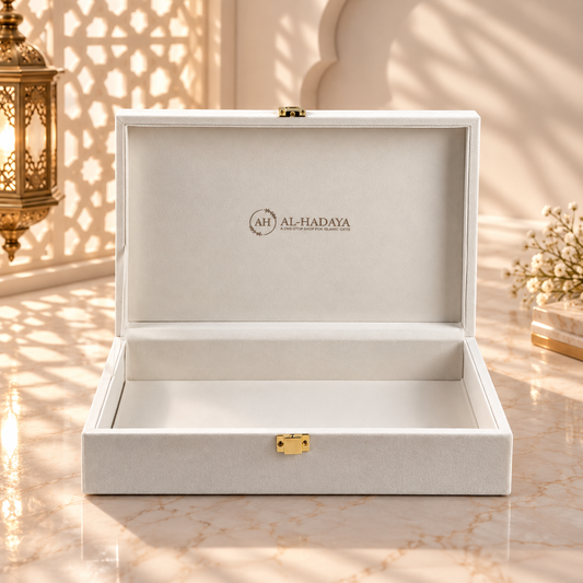 An open white luxury presentation box with a gold clasp and the brand name 'AL-HADAYA' printed inside the lid, set on a marble surface with warm sunlight and shadow patterns.