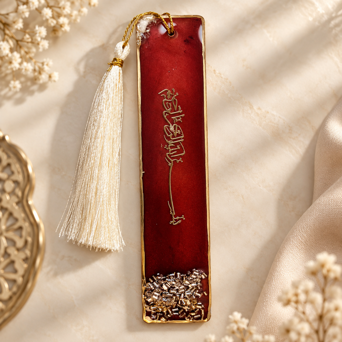 A rectangular red enamel bookmark with gold-toned metal edges, Tibetan script engraved vertically, and a cream tassel hanging from the top. The bookmark is styled on a beige fabric background with dried flowers and a gold-toned decorative object partially visible.