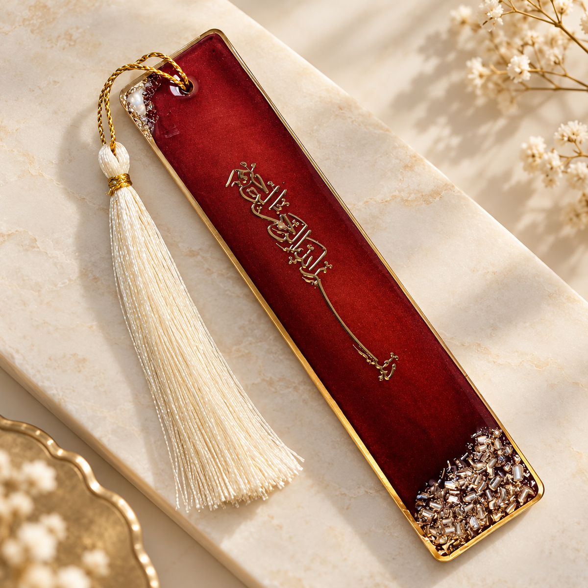 A rectangular red leather bookmark with gold trim, gold Arabic calligraphy, and a cream tassel, styled on a light marble surface with dried flowers.