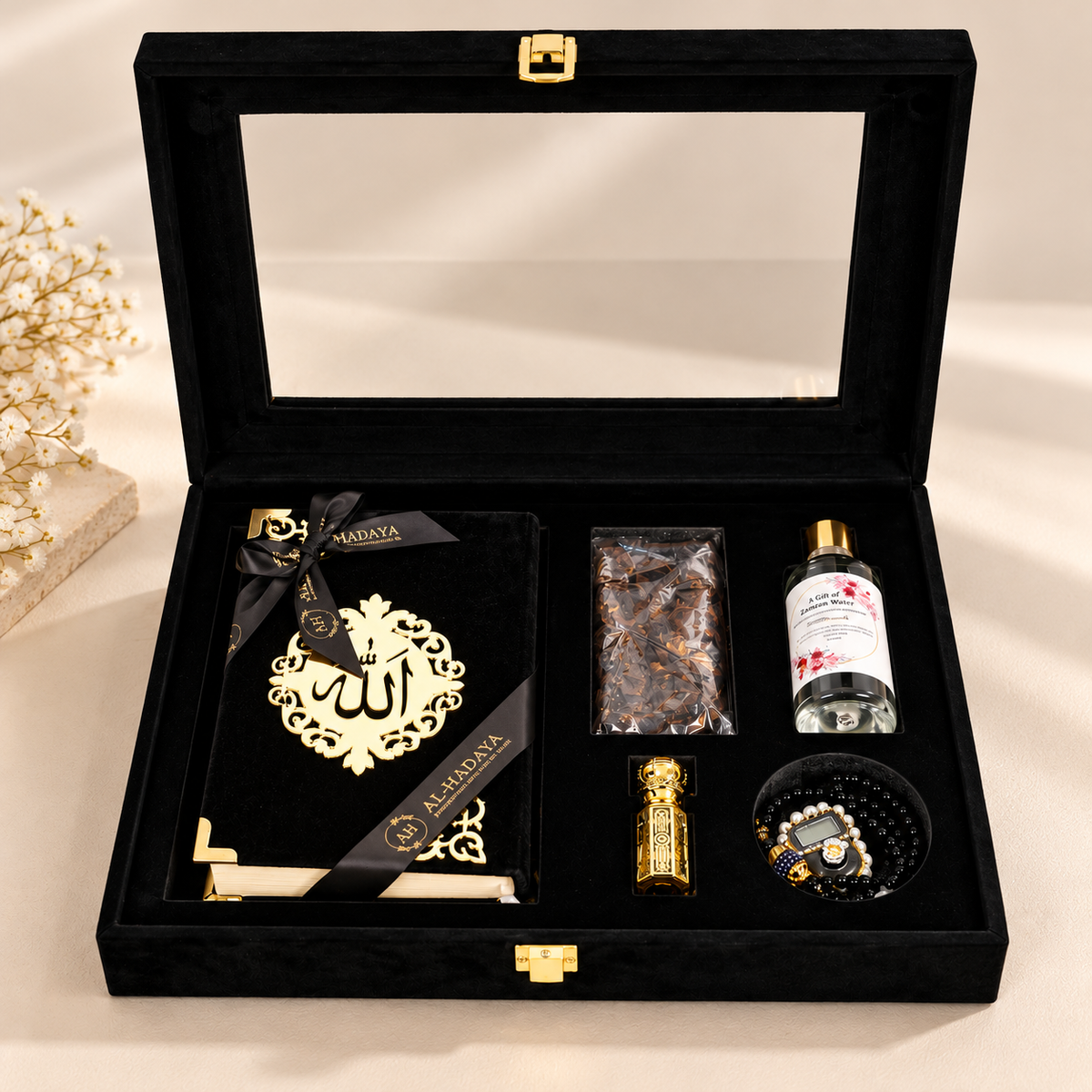 An open black velvet gift box with gold-tone hardware, arranged on a light beige surface. The box contains a black book or journal with a large gold Arabic calligraphy design and ribbon, a small gold cylindrical item, a clear bottle with a white label, a packaged brown item, and a black beaded bowl.