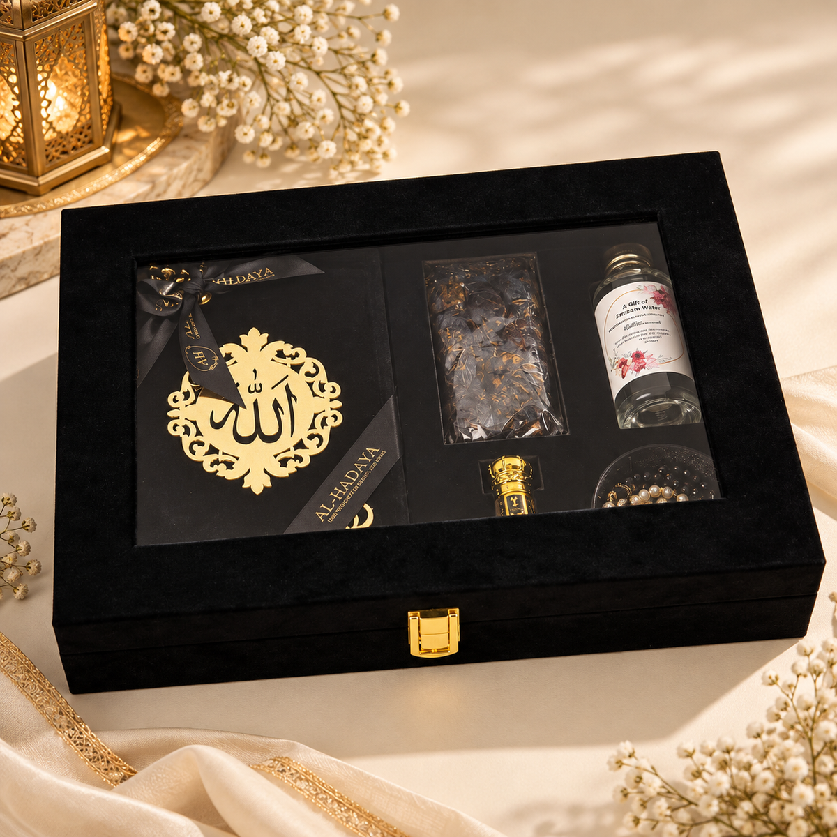A luxury black velvet gift box set with gold accents, featuring a decorative gold plaque with Arabic calligraphy, a glass bottle of rose water, and dried botanicals, styled on a cream fabric with a lantern and baby's breath.