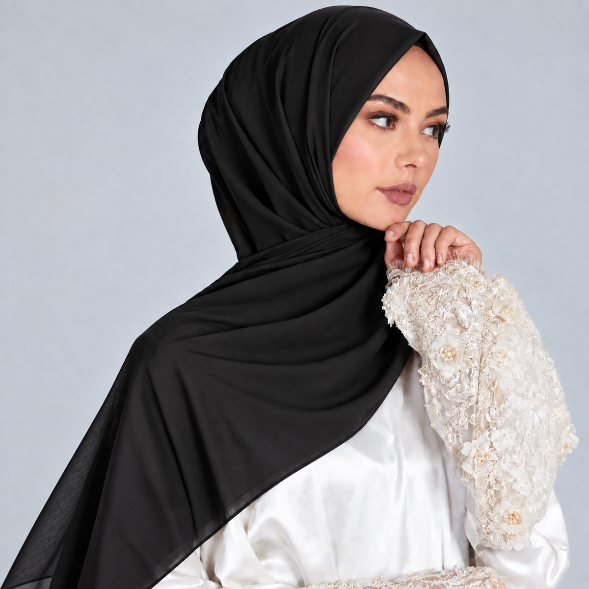 A woman wearing a black hijab draped over her shoulders and a white embellished blouse, posed against a light gray studio background.