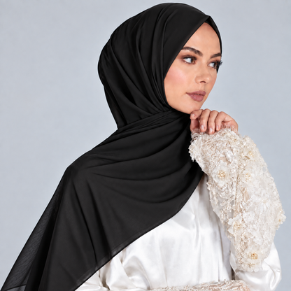 A woman wearing a black hijab draped over her shoulders and a white embellished blouse, posed against a light gray studio background.