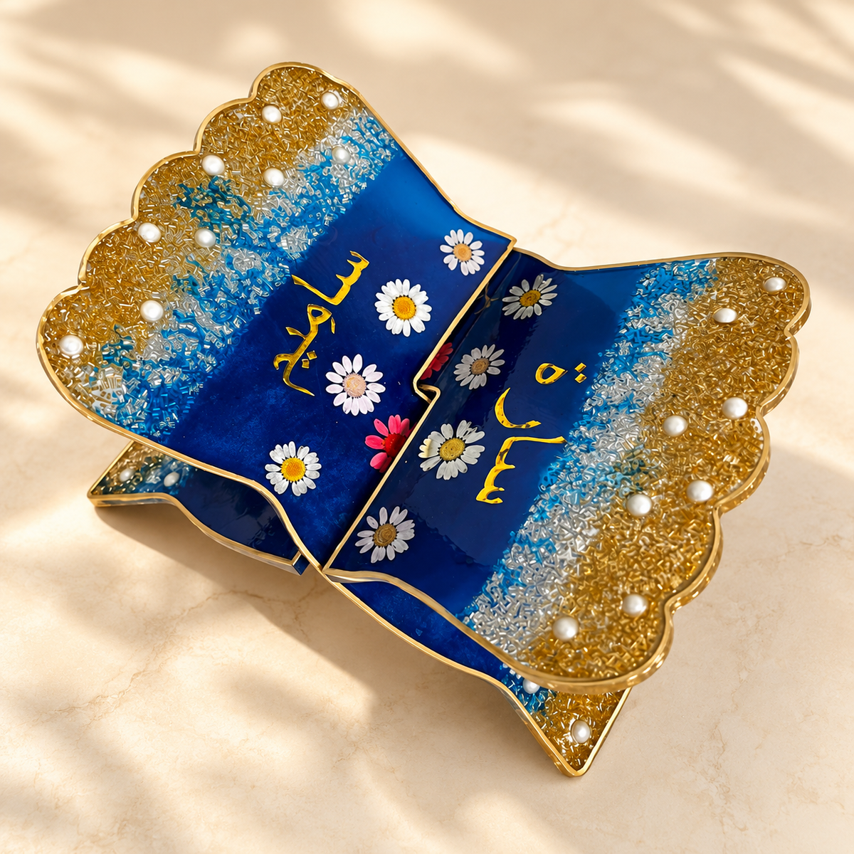 A decorative folded tray or dish with scalloped edges, featuring a deep blue center decorated with white daisy flowers and gold Arabic calligraphy, paired with glittery gold sides accented by small white pearl-like beads, photographed on a light beige surface with soft shadows.