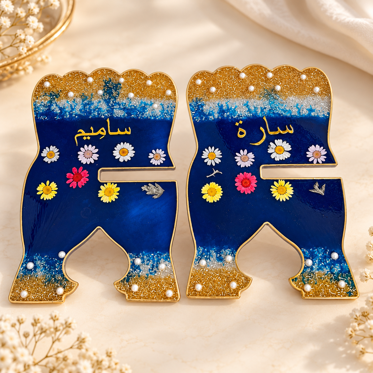 Two decorative letter ornaments shaped like legs, shown side by side on a soft cream fabric background. Each ornament has a blue center with gold glitter at the top and bottom, white pearl-like dots, small daisy flowers, and gold Arabic script.