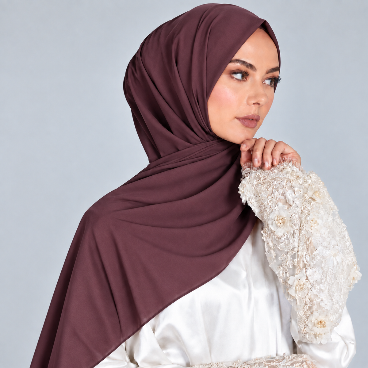 A woman wearing a draped plum-colored hijab and a white embroidered blouse, posed against a solid light gray studio background.