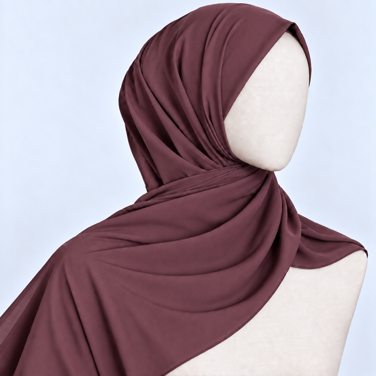 A draped hijab in a muted plum-brown color, styled on a cream-colored mannequin head and shoulders against a light blue background.