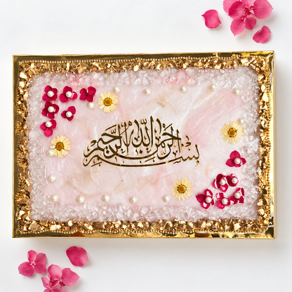 A square decorative tray or plaque with a gold metallic frame, featuring a pale pink marbled center, white pearl-like beads, small yellow daisy flowers, and a central black Arabic calligraphy design. Pink flower petals are scattered around it on a white background.