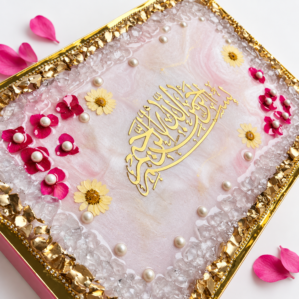 A square decorative gift box or keepsake box with a pink marbled resin top, gold foil Arabic calligraphy in the center, a gold-bezeled edge, clear crystal-like inlay, and scattered pink flower petals and small white pearl accents on a white background.