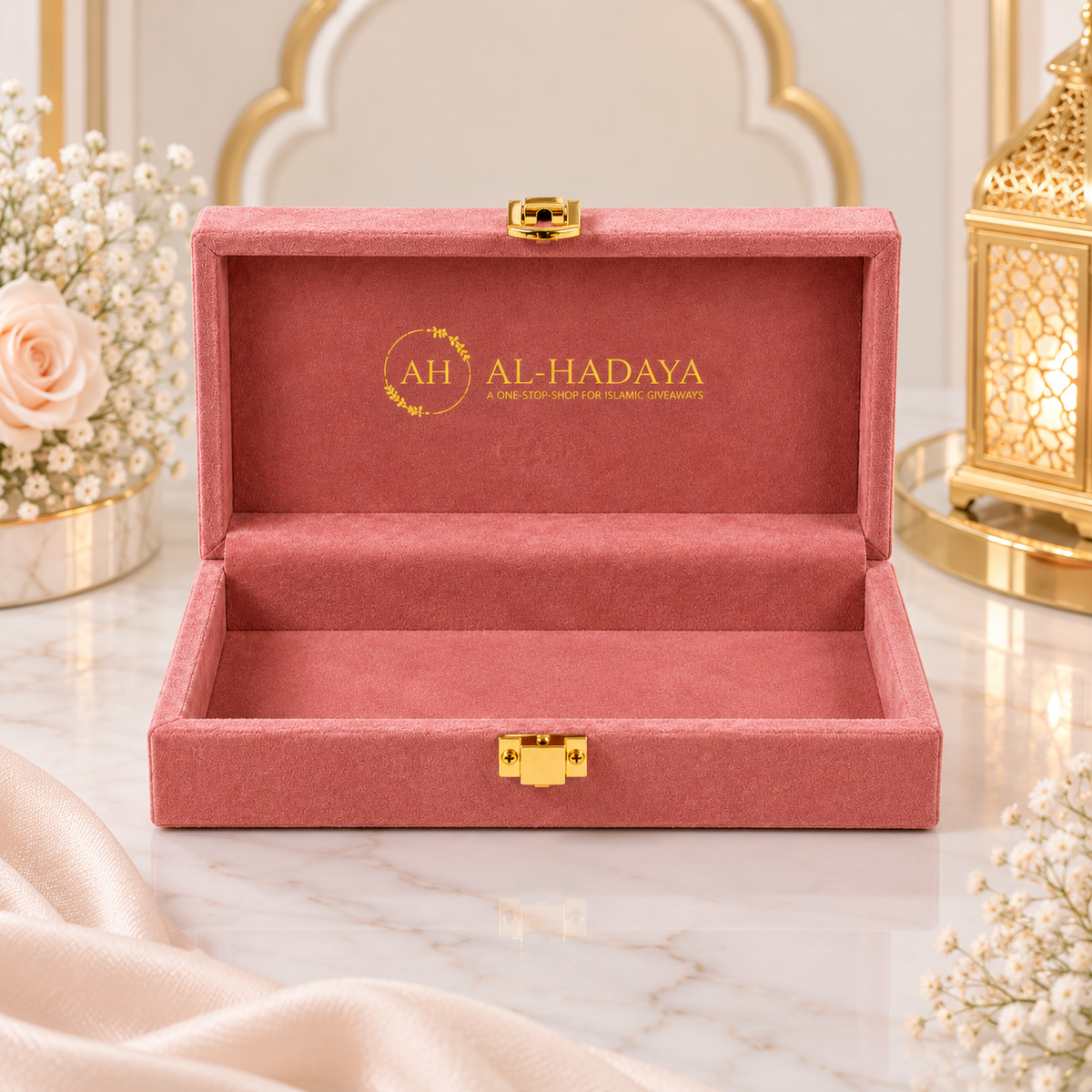 An open dusty pink velvet jewelry box with gold hardware, featuring the brand name 'AL-HADAYA' printed in gold on the inner lid, set on a marble surface with decorative flowers and a lantern.